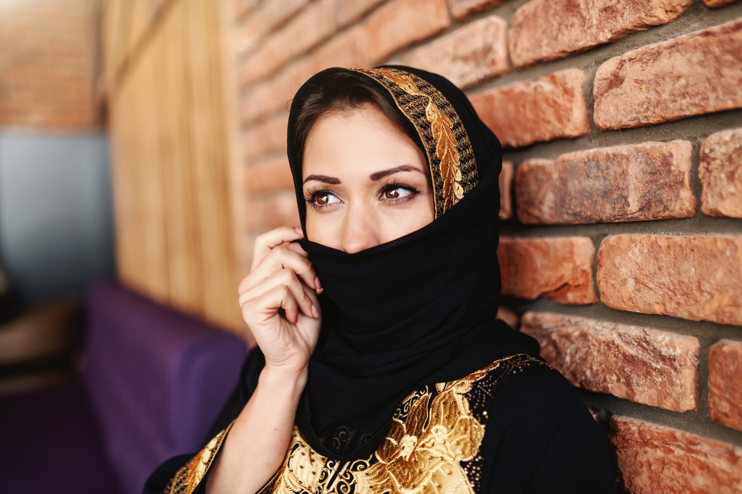Beautiful muslim woman in traditional wear covering her face with scarf while sitting in cafeteria.